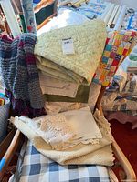 Stack on chair showing yellow quilt, crocheted throw, embroidered sham, cream knit blanket, rolled fabrics, patchwork quilt edge
