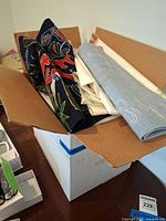 Open box showing assorted linens including dark printed fabric, light blue embroidered piece and cream cloths