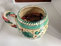 Ceramic mug decorated in green, red, yellow holding copper pennies