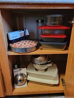 Cabinet view showing pie tin, colanders, steamer basket package, cutting boards, knife sharpener and food containers