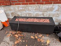 Full view of rectangular gas fire pit against wall, lava rocks visible