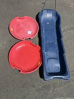 Two red saucer sleds and one blue toboggan sled arranged on pavement