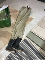Full view of khaki chest waders with integrated black boots on basement floor