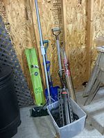 All items leaning against wall including roof rake, shovels, gutter ware box, stakes; tub with sprinkler and hose