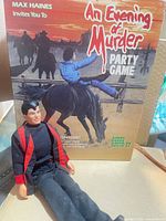 Front view of Kevin Richardson doll seated before An Evening of Murder game box