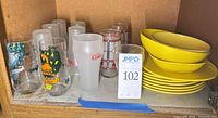 Shelf with Coca-Cola glasses, various tumblers, and stack of yellow melamine plates and bowls