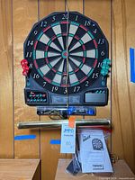 Front view of Halex Impact iS5.0 dartboard with darts, manual and brass wall light