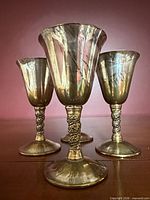 Front view of four brass cordial goblets showing ornate stems and patina