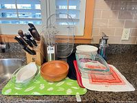 Counter view showing knife block with knives, whisks in holder, glass bake dishes, wooden bowl, mats, towels, soap dispenser