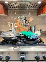 Overall lot on stove: wok with lid, stack of cast iron skillets, green oven mitts, utensil basket, stainless stock pot