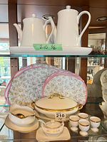 Shelved group showing two tall pots on tray, fabric covers behind, tureen, gravy boat, egg cups.