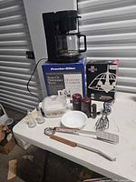 All items grouped on table including coffeemaker on box, light fixture box, casserole, utensils, mixer, shakers, burgundy containers, bowl