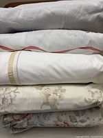Stacked view of five folded/rolled queen sheet sets showing grey flannelette, white poplin with red/blue detail, white with beige trim, cream floral, and pastel floral prints