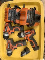 Three dusty orange Ridgid 12V drills, two chargers, one loose battery in yellow tray