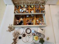 open jewelry box showing interior tray filled with assorted brooches, pins, earrings and base compartment with extra jewelry pieces