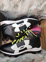 Cleats in original box showing black upper, white logo, neon yellow laces, extra laces beside