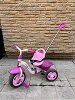 Left side view of pink and white Kettler tricycle with push handle, bucket and pedal visible