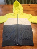 Front view of neon yellow, white, navy color block hooded windbreaker jacket laid flat showing zipper and elastic hem