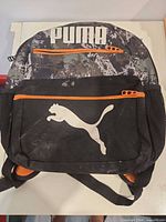 Front view showing camo top, white Puma logo, orange zipper, mesh pockets