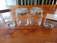 Four clear square glass jars with silver metal lids arranged largest to smallest on wooden surface
