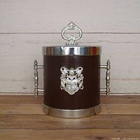 front view showing brown faux leather wrap, silver crest, lid with finial, side handles and base ring