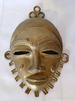 Front view of brass Ashanti mask showing facial details and hanging loops