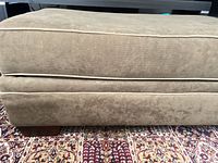 Full side view of olive green ottoman showing upholstery, piping, and wooden feet