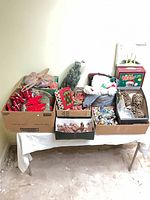 Overview of multiple boxes on table containing ornaments, bows, pine cones, mini lights and boxed Santa