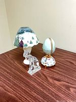 Three items: crystal cat figurine, enamel egg on stand, table lamp with painted glass shade