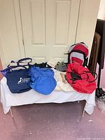 Six assorted bags laid out on table: two navy totes, blue duffel, off-white tote with purple handles, red duffel, red-grey backpack