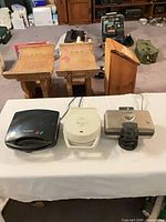 Three waffle makers lined on table: black Cuisinart, off-white Oster, stainless Proctor Silex