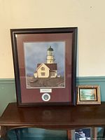 Large framed lighthouse print with medallion and small landscape painting on table