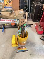Bucket containing pipe wrench, hatchet, pry bar, panel saw, hacksaw frames, floor squeegee with two yellow miter boxes beside