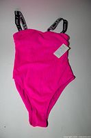 Front view of swimsuit laid flat showing color, ribbed texture, logo straps, attached tag