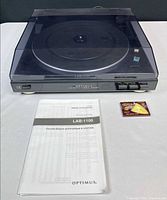 Front view of Optimus LAB-1100 turntable with dust cover closed, manual and replacement stylus visible