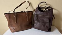 Two leather bags side by side, tan Michael Kors tote left, dark brown Fossil shoulder bag right