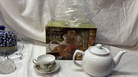 All three items: glass teapot on box, white ceramic teapot, porcelain teacup/saucer