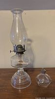 Tall clear glass oil lamp with chimney and small glass oil lamp bottle displayed on wooden surface