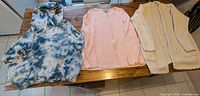 All three sweaters laid flat on table: tie-dye hoodie, pink sweater, off-white cardigan