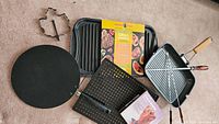 Assorted grilling items laid out: griddle tray, square pan, round pan, perforated pan, boxed roaster, maple leaf mold