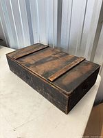 angled view of closed wooden box showing worn black/brown finish and lid cleats