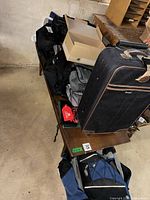 Table with rolling suitcase standing upright, multiple bags piled, blue/black backpack beneath