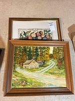 Two framed artworks stacked: landscape painting on board below, fruit print above