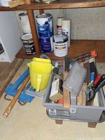 Shelf with partial paint cans, spray paints, gray tray containing hand tools, yellow pitchers and caulk guns