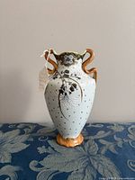 Front view of off-white porcelain vase with gilded rim and double scrolled handles, original tag visible