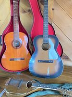 Two acoustic guitars in front of wood panel wall; left classical guitar in red case, right sunburst acoustic guitar beside it