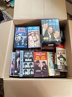 Open cardboard box showing assorted VHS tapes including Sun Valley Serenade, In Harm's Way, boxed set, Beatles tape, The Third Man, Citizen Kane
