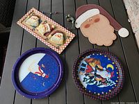 Front view of all four items: two metal plates with Christmas illustrations, wooden plaque with three Santa faces, wooden Santa head ornament