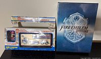 Front view of sealed Famicom controller package beside sealed Fire Emblem Warriors box