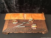 Set of three darts laid on wooden stand with open tan leather case above